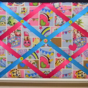 May include: A fabric memo board with a colorful Shopkins pattern. Pink and blue ribbons crisscross the board, secured by yellow buttons. The board features a variety of Shopkins characters and a business card.