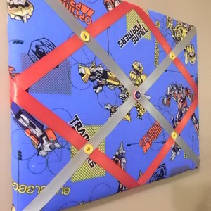 May include: A blue fabric memo board featuring Transformers characters. The board has red and silver ribbons crisscrossing the surface, secured with yellow buttons. The words "TRANSFORMERS" and "OPTIMUS PRIME" are visible on the fabric.