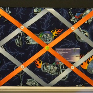 May include: A rectangular memo board with a dark blue fabric background featuring a motorcycle and lightning bolt pattern. The board has a crisscross design of silver and orange ribbons, secured with white and yellow buttons. A business card is visible.