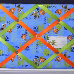 May include: A fabric memo board featuring a blue background with Nemo and Squirt characters, green leaves, and anchor patterns. The board has orange and green ribbons crisscrossing the surface, secured with gray buttons. A business card is visible.