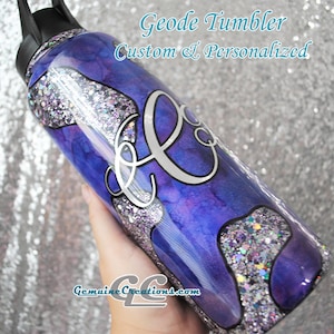 Peut inclure: Un gobelet cylindrique avec un couvercle noir présentant une finition marbrée violette et bleue avec des accents de paillettes argentées. La surface affiche un grand monogramme en écriture blanche et le texte Geode Tumbler Custom & Personalized et GenuineCreations.com.