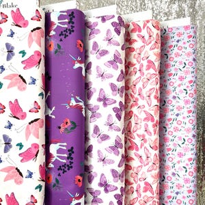 Peut inclure: Cinq rouleaux de tissu verticaux disposés côte à côte. Le premier présente des papillons roses et violets sur fond blanc. Le deuxième est violet avec des licornes et des fleurs. Le troisième a des papillons violets sur blanc. Le quatrième montre des papillons roses sur blanc. Le cinquième a des fleurs roses et violettes sur blanc.