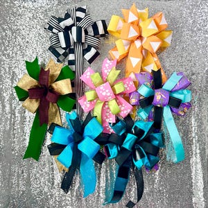 May include: An assortment of decorative bows in various colors and patterns. The bows include black and white stripes, orange and white triangles, pink with white hearts, and teal with black accents. The bows are arranged on a sparkly silver background.