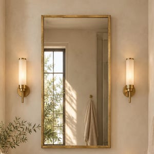 Peut inclure: Un miroir mural rectangulaire avec un cadre doré fin, encadré par deux appliques cylindriques en verre dépoli avec des fixations dorées. L'ensemble comprend un comptoir en marbre, un robinet doré, un vase en céramique et une bougie allumée.