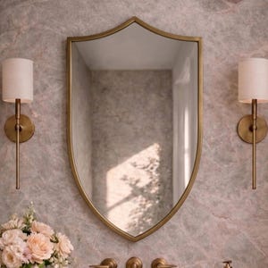 Puede incluir: Un espejo dorado en forma de escudo cuelga sobre una encimera de mármol con un lavabo y grifos dorados. Dos apliques con pantallas blancas flanquean el espejo. Un jarrón de flores y una bandeja con artículos de tocador completan la escena.