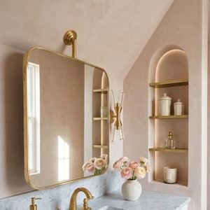 May include: A bathroom vanity with a marble countertop, white cabinets, and gold fixtures. A gold-framed mirror hangs above the sink, reflecting the room. A vase of flowers sits on the counter, and shelves with decorative items are built into the wall.