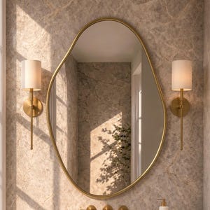 Puede incluir: Un espejo de pared ovalado irregular con un marco dorado fino, flanqueado por dos apliques de pared verticales con pantallas blancas cilíndricas. Debajo, un grifo dorado montado sobre una encimera de mármol con una bandeja y jabonera.