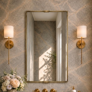 May include: A rectangular wall mirror with a thin brass frame centered between two brass wall sconces with white cylindrical shades. Below the mirror is a stone countertop with a built-in sink, brass faucet handles, a floral vase, and a tray.