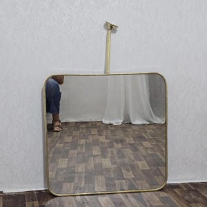 May include: A square, gold-framed mirror with rounded corners hangs from a gold rod. The mirror reflects a room with a white curtain and a person wearing blue jeans and sandals. The mirror is hanging on a white wall.