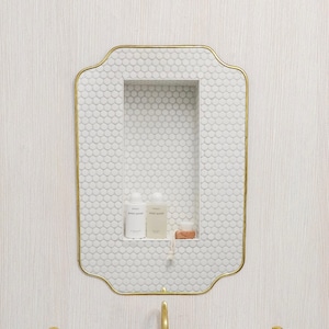 May include: A bathroom sink with a copper basin and a turquoise exterior. The sink is set on a white cabinet, with a gold faucet and two gold soap dispensers. Above the sink is a white tiled niche with a gold frame.