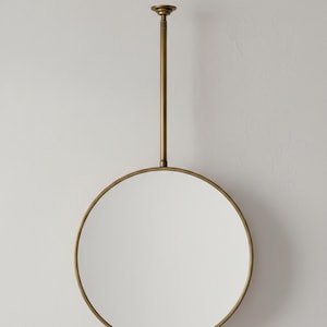 May include: A circular hanging mirror with a brass-toned frame and a matching rod. The mirror has a reflective surface. The rod extends from the top of the mirror to a ceiling mount. The design is simple and elegant, suitable for various interior styles.