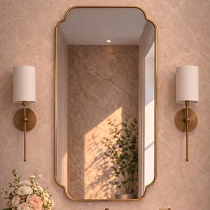 Op de afbeelding: Badkamerinterieur met een goudomrand spiegel, gouden wandlampen met witte kappen en een marmeren oppervlak. Een vaas met bloemen, een zeepdispenser en handdoeken zijn te zien. De muur heeft een lichte, patroonafwerking.