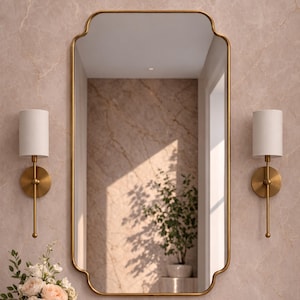 May include: A rectangular mirror with a gold frame and a decorative top is mounted above a bathroom sink. Two wall sconces with white shades are either side of the mirror. The bathroom features gold fixtures and a vase of flowers.