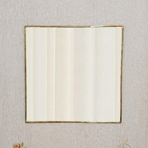 Può includere: Uno specchio rettangolare con cornice dorata è appeso a una parete strutturata grigio chiaro. La superficie dello specchio riflette strisce verticali di luce e bianco sporco. Un piccolo vaso dorato con fiori rosa e una pianta decorativa sono sotto.