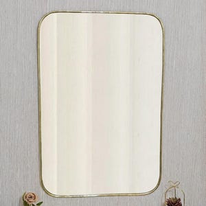 May include: Rectangular mirror with rounded corners and a gold-colored frame. The mirror is mounted on a light-colored wooden surface, with a small gold-colored pot of flowers and a decorative plant on either side.