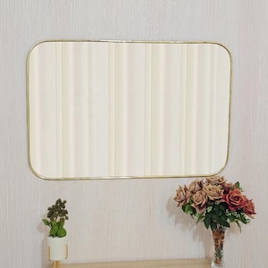 May include: Rectangular mirror with a gold-colored frame hanging on a textured wall. Below the mirror is a wooden cabinet with a small plant in a white pot and a vase of roses.