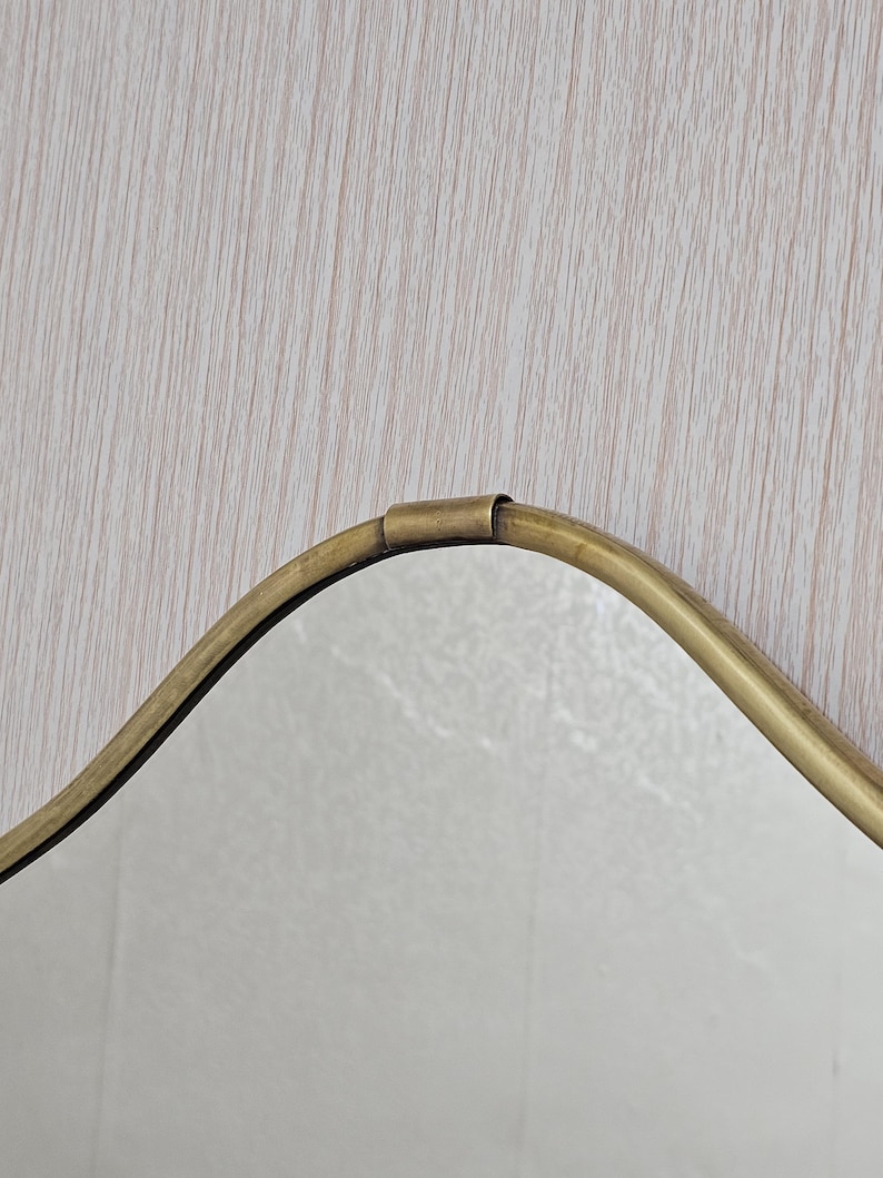K&ouml;nnte beinhalten: Ein Spiegel im Vintage-Stil mit einer wellenf&ouml;rmigen, unregelm&auml;&szlig;igen Form. Der Rahmen besteht aus goldfarbenem Metall, und die Oberfl&auml;che des Spiegels ist reflektierend. Das Design ist schlicht und elegant, passend f&uuml;r verschiedene Einrichtungsstile.