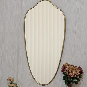 K&ouml;nnte beinhalten: Ein ovaler Spiegel mit cremefarbenem, vertikal gestreiftem Mittelteil und einem goldfarbenen Rahmen. Der Spiegel h&auml;ngt an einer grauen Wand, mit einer kleinen goldenen Vase mit rosa Blumen und einer Vase mit Rosen.
