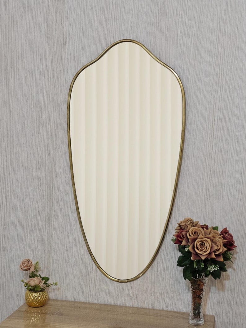 K&ouml;nnte beinhalten: Ein ovaler Spiegel mit cremefarbenem, vertikal gestreiftem Mittelteil und einem goldfarbenen Rahmen. Der Spiegel h&auml;ngt an einer grauen Wand, mit einer kleinen goldenen Vase mit rosa Blumen und einer Vase mit Rosen.