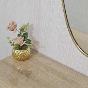K&ouml;nnte beinhalten: Ein kleiner, goldfarbener, strukturierter dekorativer Topf mit k&uuml;nstlichen rosa Rosen und Gr&uuml;nzeug. Der Topf steht auf einer hellen Holzoberfl&auml;che, mit einem goldgerahmten ovalen Spiegel im Hintergrund.