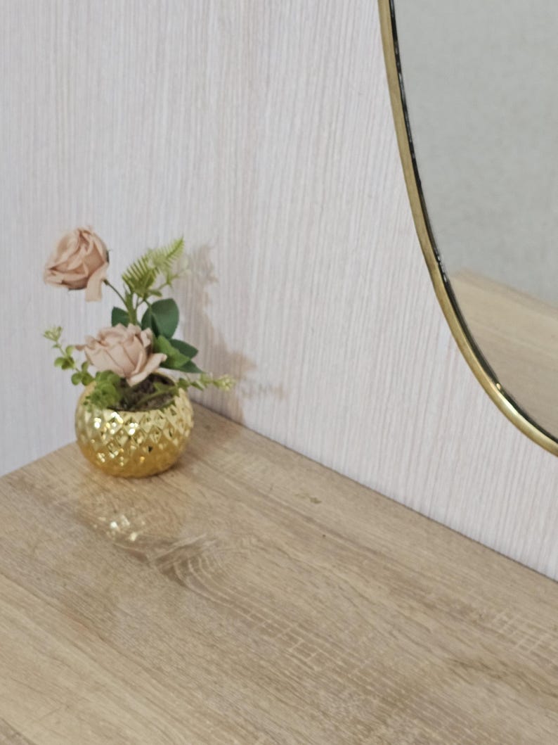 K&ouml;nnte beinhalten: Ein kleiner, goldfarbener, strukturierter dekorativer Topf mit k&uuml;nstlichen rosa Rosen und Gr&uuml;nzeug. Der Topf steht auf einer hellen Holzoberfl&auml;che, mit einem goldgerahmten ovalen Spiegel im Hintergrund.