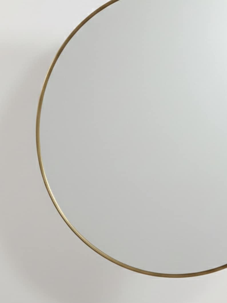 May include: A circular mirror featuring a slender, gold-toned metal frame. The reflective surface of the mirror occupies the majority of the frame, reflecting the surroundings. The design is minimalist and contemporary, fitting diverse interior aesthetics.