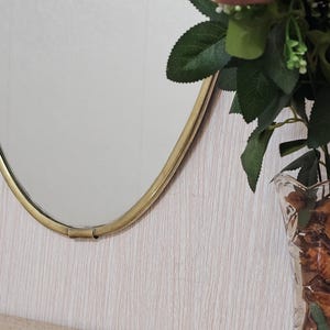 K&ouml;nnte beinhalten: Ein ovaler Spiegel mit goldenem Rahmen ist an einer hellen, strukturierten Wand befestigt. Rechts vom Spiegel steht eine klare Glasvase mit getrockneten Blumen und Gr&uuml;n. Die Vase steht auf einer Holzoberfl&auml;che.