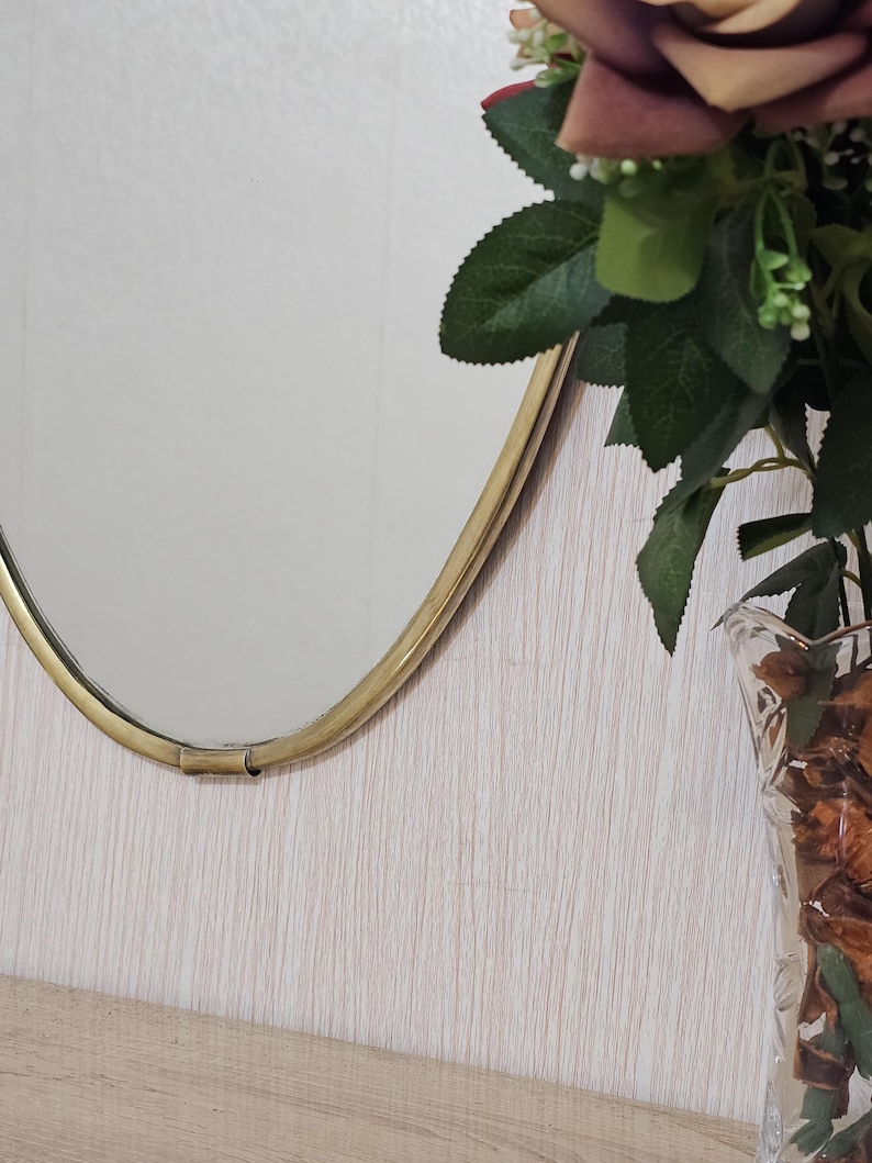 K&ouml;nnte beinhalten: Ein ovaler Spiegel mit goldenem Rahmen ist an einer hellen, strukturierten Wand befestigt. Rechts vom Spiegel steht eine klare Glasvase mit getrockneten Blumen und Gr&uuml;n. Die Vase steht auf einer Holzoberfl&auml;che.