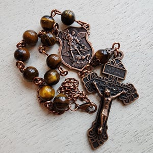 May include: A copper toned rosary with tiger's eye beads, a crucifix with the inscription "JESUS NAZARENUS REX JUDAEORUM", and a Saint Michael medal.