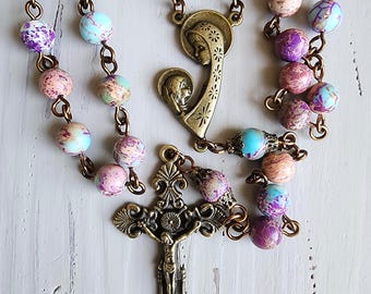 May include: A rosary featuring round, multi-colored jasper beads in shades of purple, blue, and pink, connected by bronze-toned metal links. The piece includes a central bronze-toned medal depicting two figures and a detailed bronze-toned crucifix.