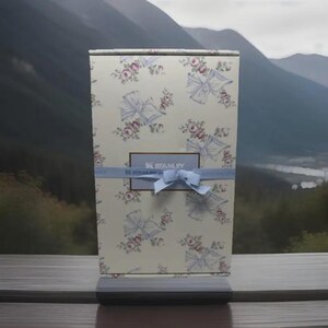 Op de afbeelding: Een rechthoekige geschenkdoos met een bloemen- en strikpatroon in roze en blauw. De doos is versierd met een lichtblauw lint en een strik. De merknaam "STANLEY" is zichtbaar op de doos.