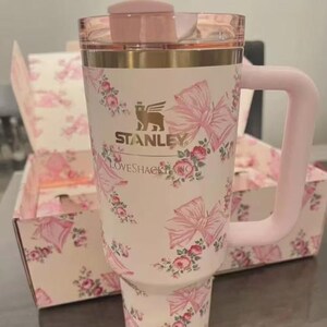 May include: A white Stanley travel mug with a pink handle, lid, and trim. The mug features a pattern of pink bows and roses. The matching box in the background has the same design. The text "STANLEY" and "LOVESHACKFANCY" are printed on the mug.