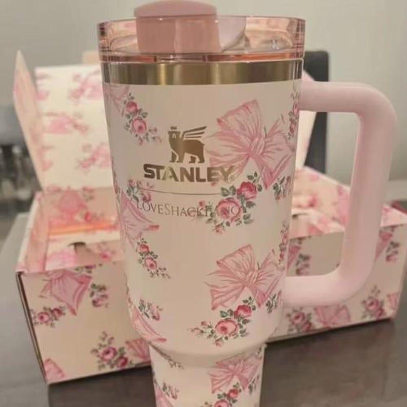 May include: A white Stanley travel mug with a pink handle, lid, and trim. The mug features a pattern of pink bows and roses. The matching box in the background has the same design. The text "STANLEY" and "LOVESHACKFANCY" are printed on the mug.