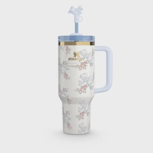 Op de afbeelding: Een witte Stanley-beker met een bloemen- en strikpatroon. De beker heeft een lichtblauwe handgreep, deksel en rietje met een strikvormige top. De tekst "Stanley" en "LoveShackFancy" zijn gedrukt.