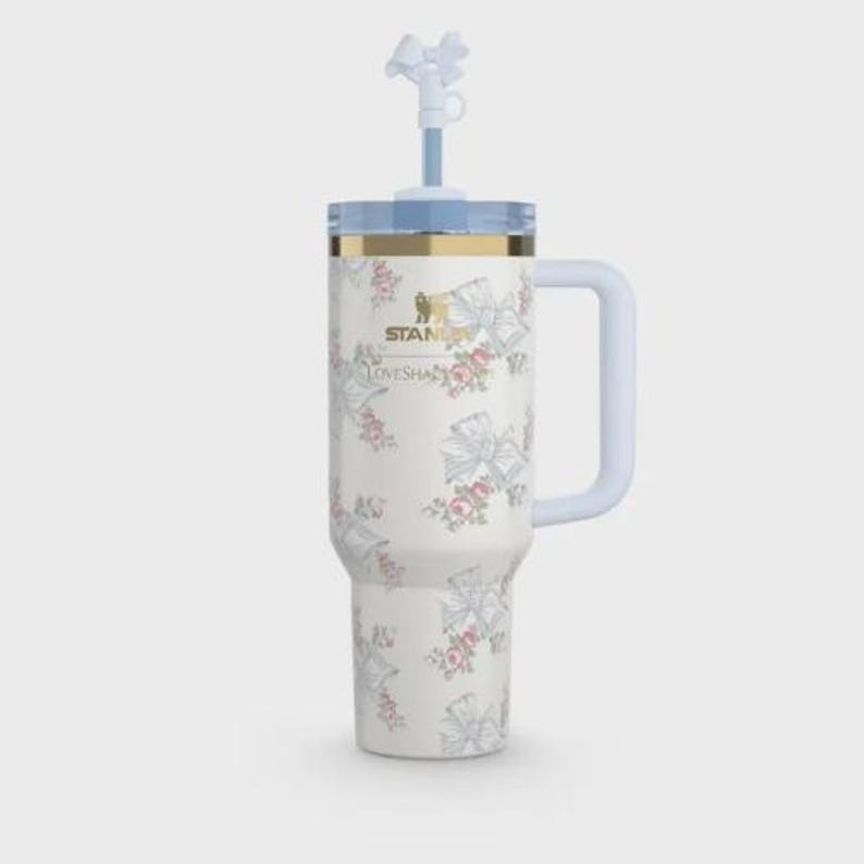 Op de afbeelding: Een witte Stanley-beker met een bloemen- en strikpatroon. De beker heeft een lichtblauwe handgreep, deksel en rietje met een strikvormige top. De tekst "Stanley" en "LoveShackFancy" zijn gedrukt.
