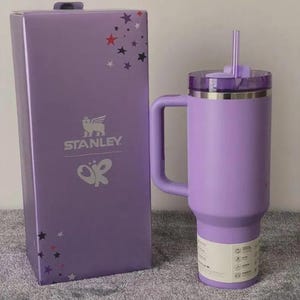 May include: A lavender Stanley tumbler with a handle and straw, next to a matching box. The box features the Stanley logo, a butterfly design, and star accents. The tumbler has a capacity of approximately 40 ounces.
