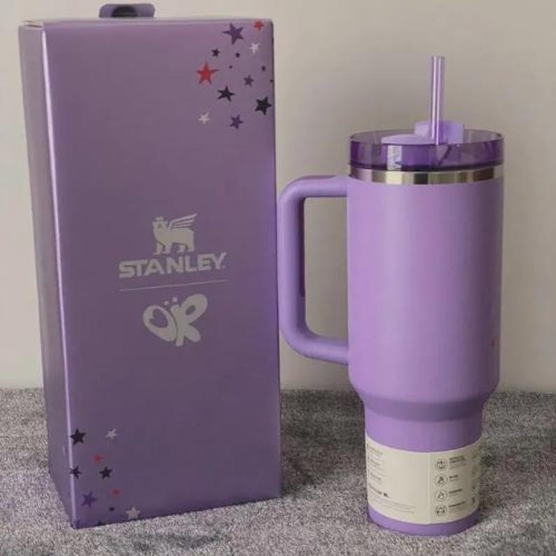 May include: A lavender Stanley tumbler with a handle and straw, next to a matching box. The box features the Stanley logo, a butterfly design, and star accents. The tumbler has a capacity of approximately 40 ounces.
