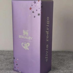 May include: A tall, rectangular box in a light purple hue, adorned with silver stars and the "Stanley" logo. The text "olivia rodrigo" is printed vertically on the side. A butterfly graphic is also visible.