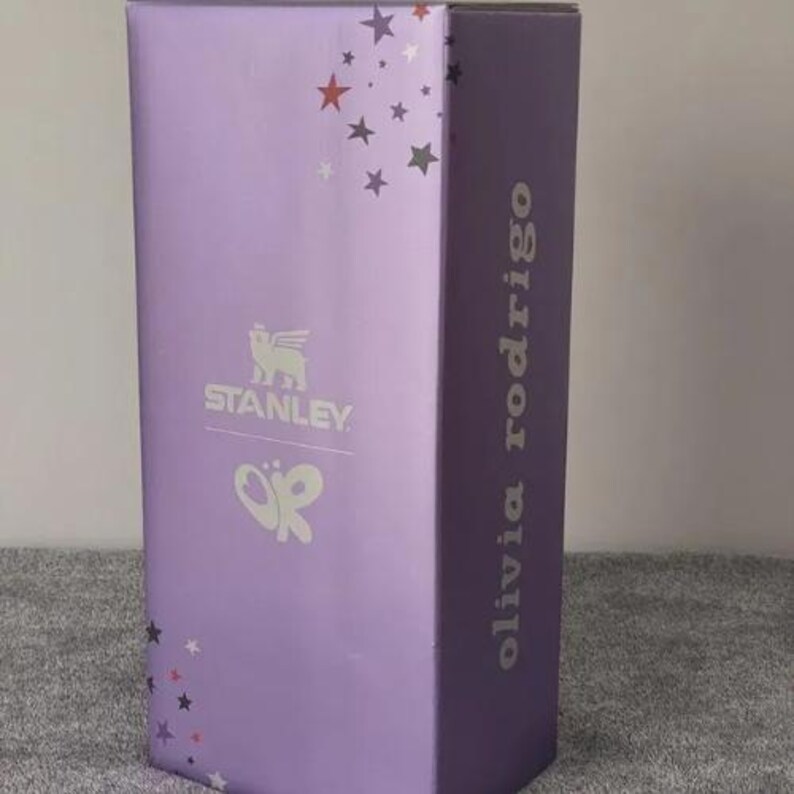 May include: A tall, rectangular box in a light purple hue, adorned with silver stars and the "Stanley" logo. The text "olivia rodrigo" is printed vertically on the side. A butterfly graphic is also visible.