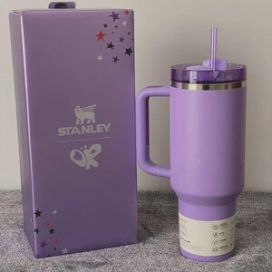 Peut inclure: Un gobelet Stanley lavande avec une poign&eacute;e et une paille, &agrave; c&ocirc;t&eacute; d'une bo&icirc;te assortie. La bo&icirc;te pr&eacute;sente le logo Stanley et des &eacute;toiles. Le gobelet a un couvercle transparent et une &eacute;tiquette blanche.