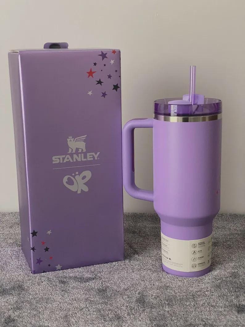 Peut inclure: Un gobelet Stanley lavande avec une poign&eacute;e et une paille, &agrave; c&ocirc;t&eacute; d'une bo&icirc;te assortie. La bo&icirc;te pr&eacute;sente le logo Stanley et des &eacute;toiles. Le gobelet a un couvercle transparent et une &eacute;tiquette blanche.