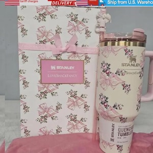 May include: A Stanley Quencher tumbler and gift box, decorated with a pink and cream floral and bow design. The tumbler has a handle and a lid. The gift box features the Stanley and LoveShackFancy logos.