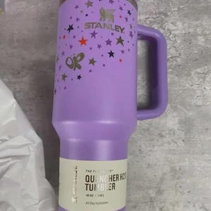 May include: A purple Stanley Quencher H2.0 Tumbler with a handle, decorated with stars. The tumbler has the Stanley logo and the text "OR" on the side. The label reads "QUENCHER H2.0 TUMBLER" and "40 OZ | 1.18 L".
