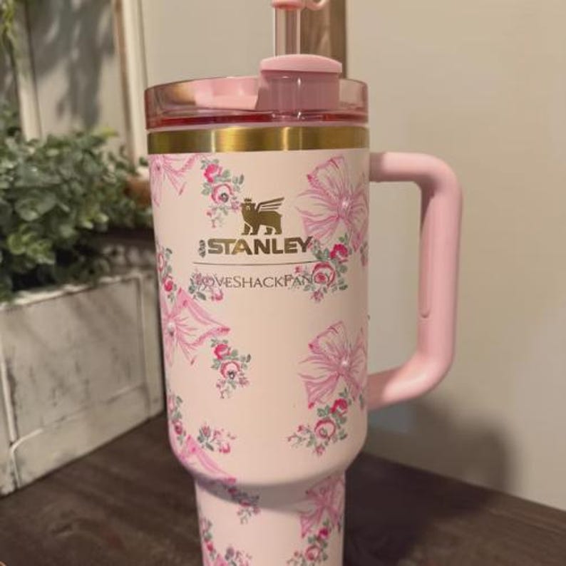 May include: A pink Stanley travel mug with a floral pattern. The mug features a pink handle, lid, and trim. The Stanley logo and "LOVESHACKFANCY" are visible. The mug is designed to hold approximately 1.18 liters.