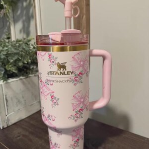 May include: A pink Stanley travel mug with a floral pattern. It features a handle, a clear lid, and a pink straw. The Stanley logo and the text "GROOVESHACKFAN" are printed on the mug.