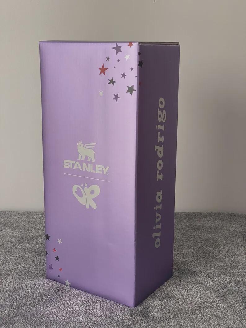 Peut inclure: Une bo&icirc;te rectangulaire haute de couleur violet clair. La bo&icirc;te pr&eacute;sente le logo "STANLEY" et le nom "olivia rodrigo" en lettres argent&eacute;es. Des &eacute;toiles d&eacute;coratives rouges, grises et blanches ornent la bo&icirc;te.
