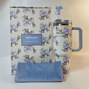Op de afbeelding: Een witte Stanley-beker met een blauw strik- en bloemenpatroon, een bijpassende doos en een blauwe doek. De beker heeft een handvat en een rietje. Op de doos staat "STANLEY LOVESHACKFANCY".