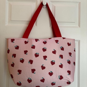 May include: A pink tote bag with red handles and a pattern of red hearts with green stems.