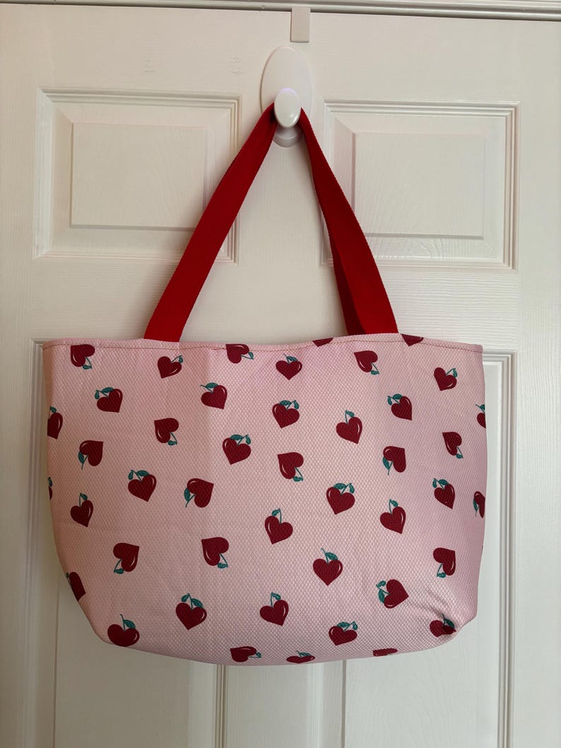 May include: A pink tote bag with red handles and a pattern of red hearts with green stems.
