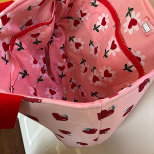 May include: Pink tote bag with a red and white cherry print and a red and white floral print lining. The bag has a red zippered pocket on the inside.